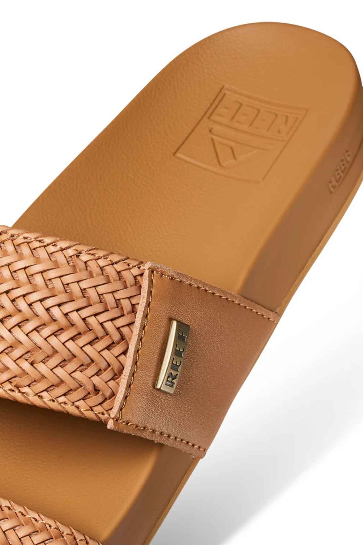 Close-up of a brown REEF Vista Braid II sandal with a woven strap, branded metal tag, cushioned midsole, and smooth textured sole. Crafted from eco-friendly materials, it features an embossed logo.
