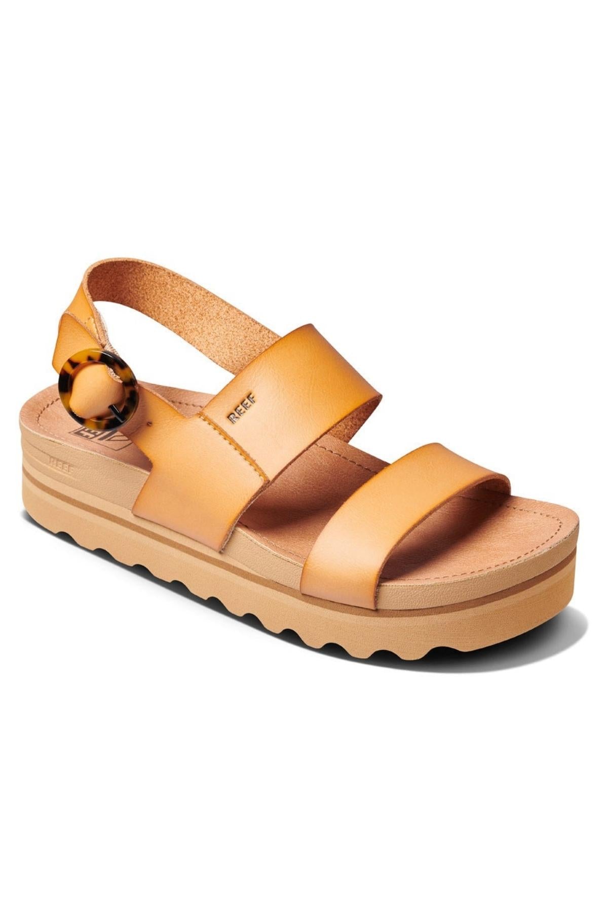 The REEF Vista Hi Buckle sandals are tan, open-toe sandals featuring two wide straps across the foot and a slingback strap with a buckle closure. They come with a vegan leather upper, a platform sole with a ridged bottom for added traction, and arch support for all-day wear.