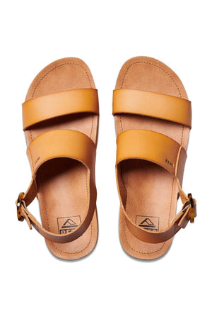 A pair of REEF Vista Hi Buckle sandals in tan, featuring two horizontal straps and a buckle ankle strap, is shown against a white background. These sandals have cushioned footbeds and arch support, with a small logo near the heel. The upper is made of vegan leather.
