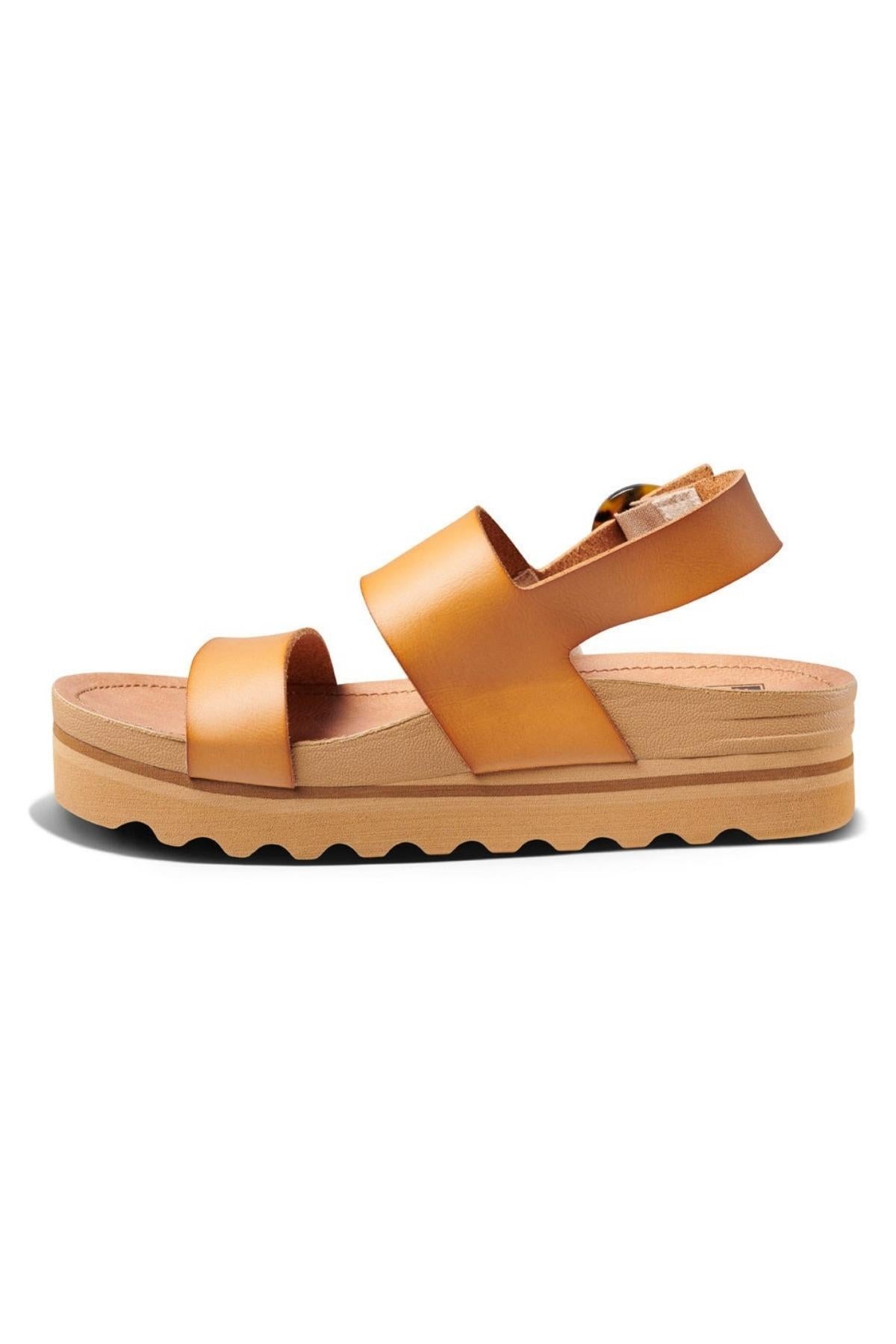 A side view of the REEF Vista Hi Buckle sandals in tan showcases the vegan leather upper with two wide straps across the foot and a buckle strap around the ankle. The sandal also features a thick, wavy-edged sole and excellent arch support.