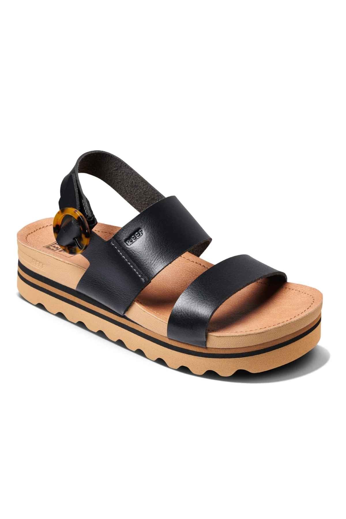 The REEF Vista Hi Buckle is a black vegan leather sandal featuring two wide straps, an adjustable heel buckle, a tan cushioned sole, and a wavy black outsole.