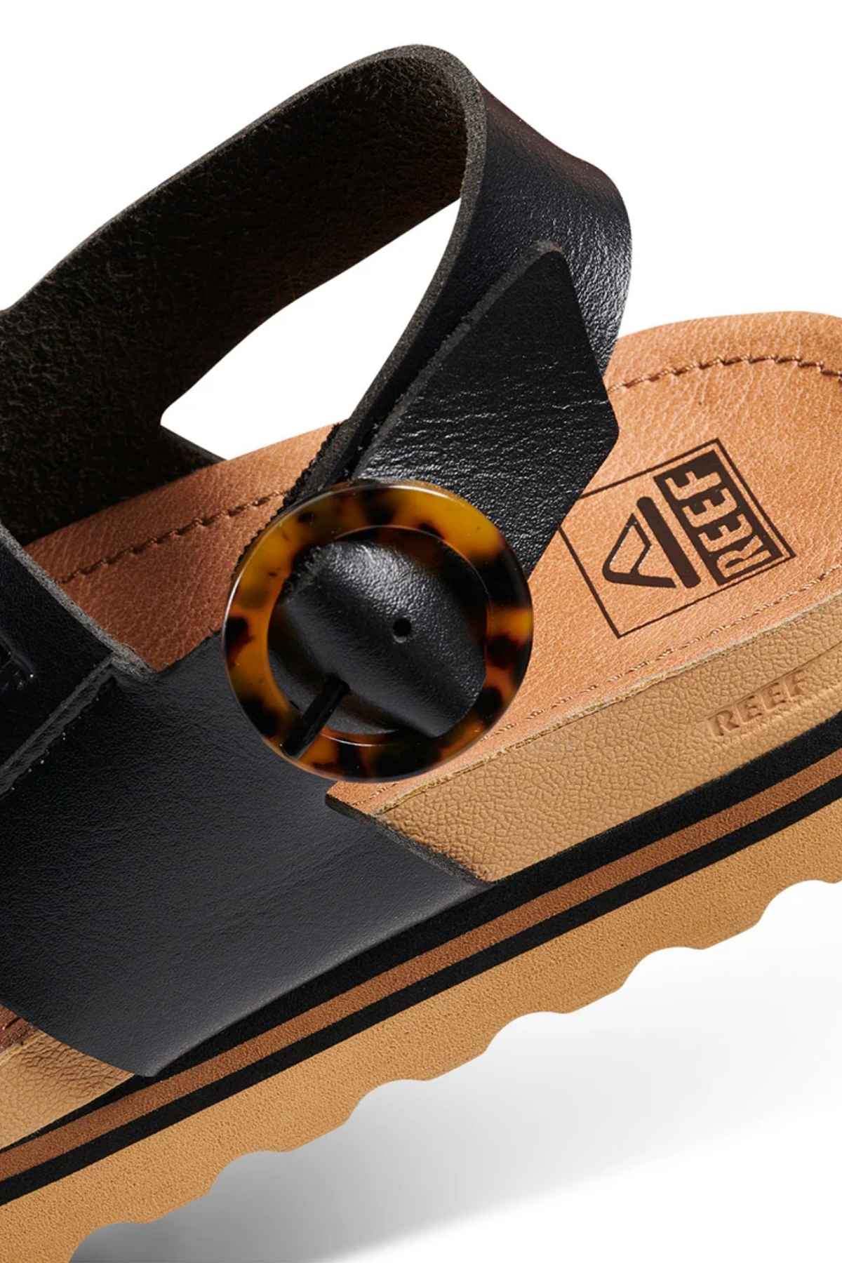Close-up of the REEF Vista Hi Buckle sandal featuring a black vegan leather strap with a tortoiseshell buckle, tan footbed embossed with the REEF logo, and a textured tan and black sole—ideal for platform sandal lovers.