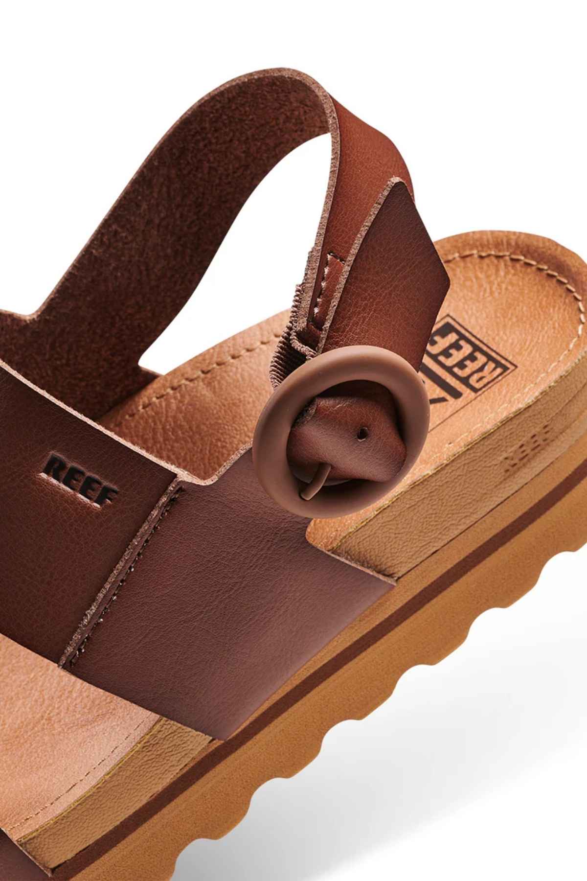 A detailed view of the REEF Vista Hi Buckle sandal featuring brown vegan leather straps, a circular buckle, and a tan platform sole. The sandal showcases the "REEF" branding on both the strap and footbed, with a wavy textured sole.