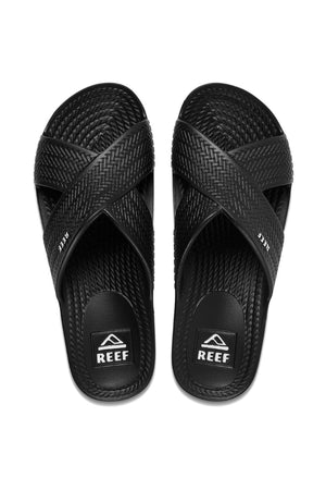 Black REEF Water X Slide sandals feature crisscross textured straps and a non-marking sugarcane EVA footbed. The soles have a textured finish, with "REEF" branded on the footbed. They are displayed side by side on a white background.