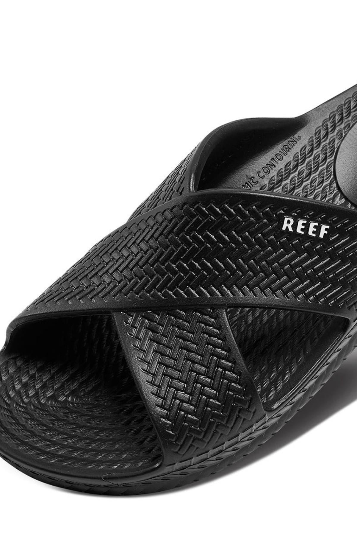 Close-up of the REEF Water X Slide sandal in black with a crisscross design and visible "REEF" logo. It features an open-toe style, contoured comfort with a non-marking sugarcane EVA footbed, highlighting eco-friendly innovation.