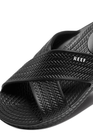 Close-up of the REEF Water X Slide sandal in black with a crisscross design and visible "REEF" logo. It features an open-toe style, contoured comfort with a non-marking sugarcane EVA footbed, highlighting eco-friendly innovation.