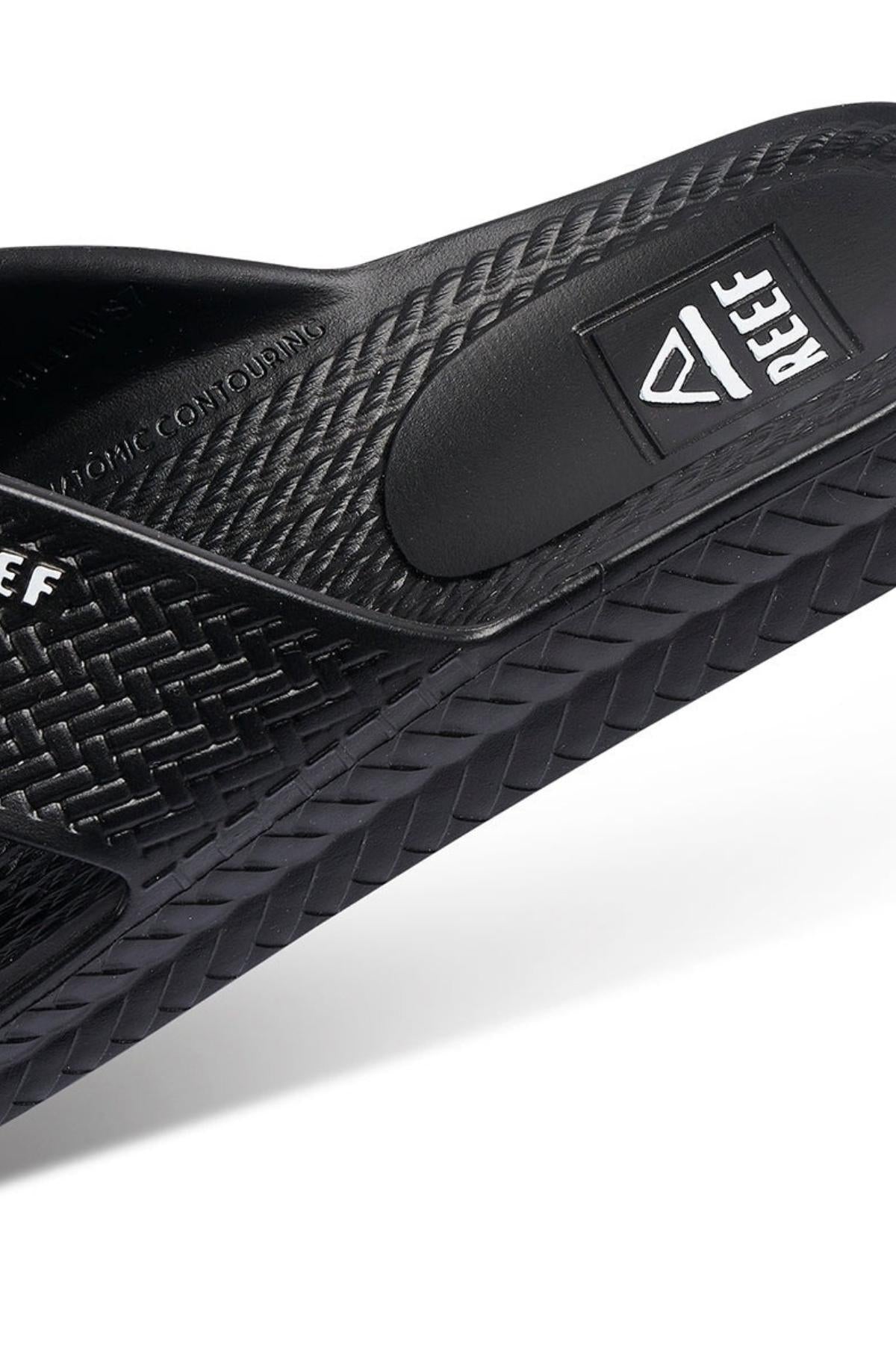 Close-up of the REEF Water X Slide in black, highlighting the "REEF" logo. It has Injection Molded Waterproof EVA construction and a non-marking sugarcane EVA footbed, with a sole featuring a woven-like pattern.