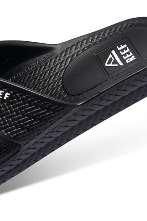 Close-up of the REEF Water X Slide in black, highlighting the "REEF" logo. It has Injection Molded Waterproof EVA construction and a non-marking sugarcane EVA footbed, with a sole featuring a woven-like pattern.