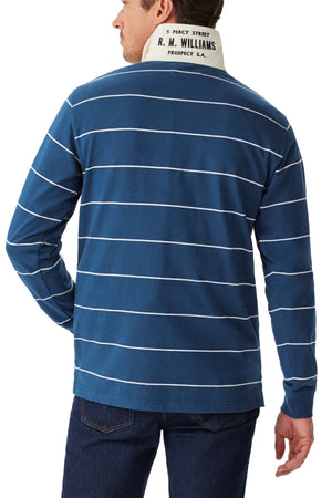 A man wearing the RM Williams Albany Stripe Rugby—a blue long-sleeve shirt with white horizontal stripes, a white collar bearing "R. M. WILLIAMS" and an address—paired with dark jeans, is facing away from the camera.
