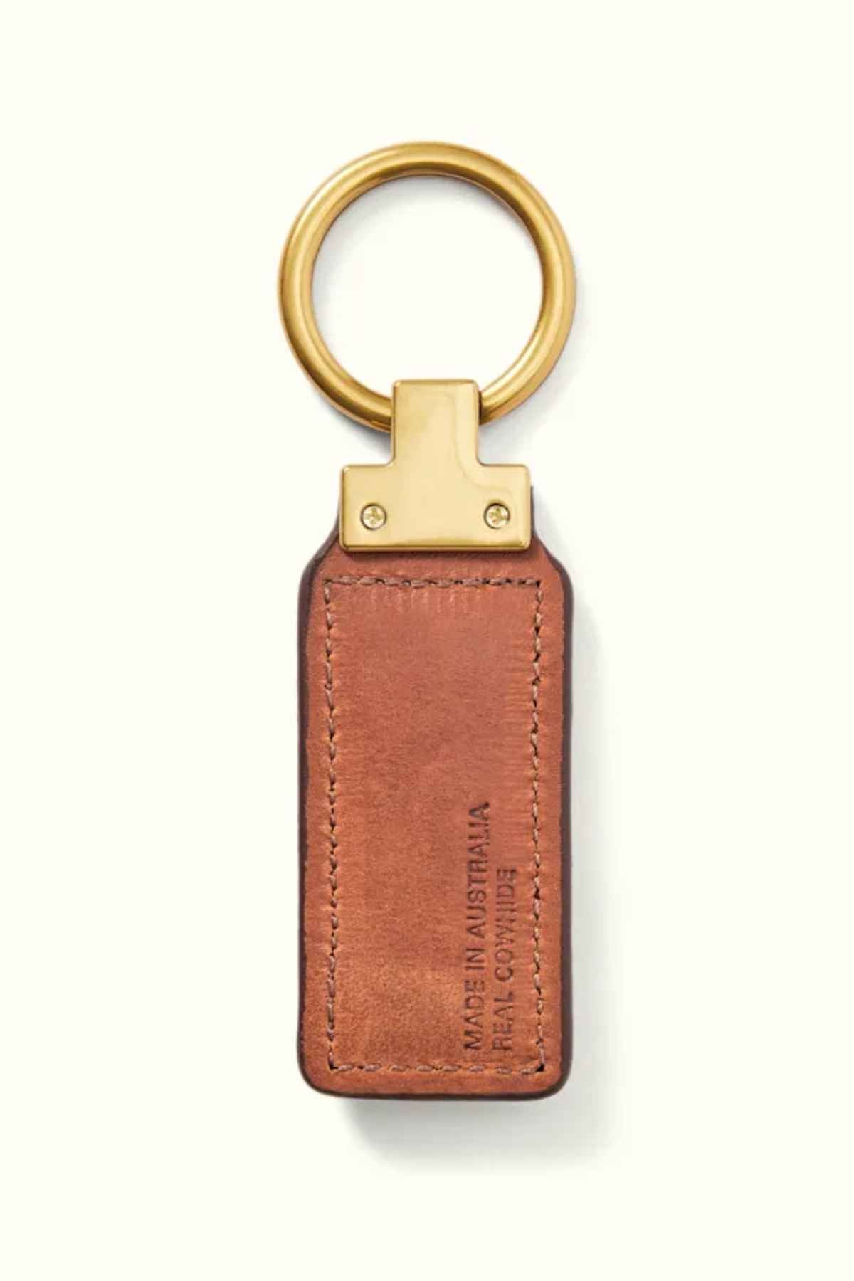The RM Williams Angaston Key Fob is a brown full-grain leather keychain featuring antique brass hardware, complemented by a gold metal ring and attachment. The rectangular leather piece is embossed with "Made in Australia R.M.W. Comfort," maintaining a simple and elegant style throughout.