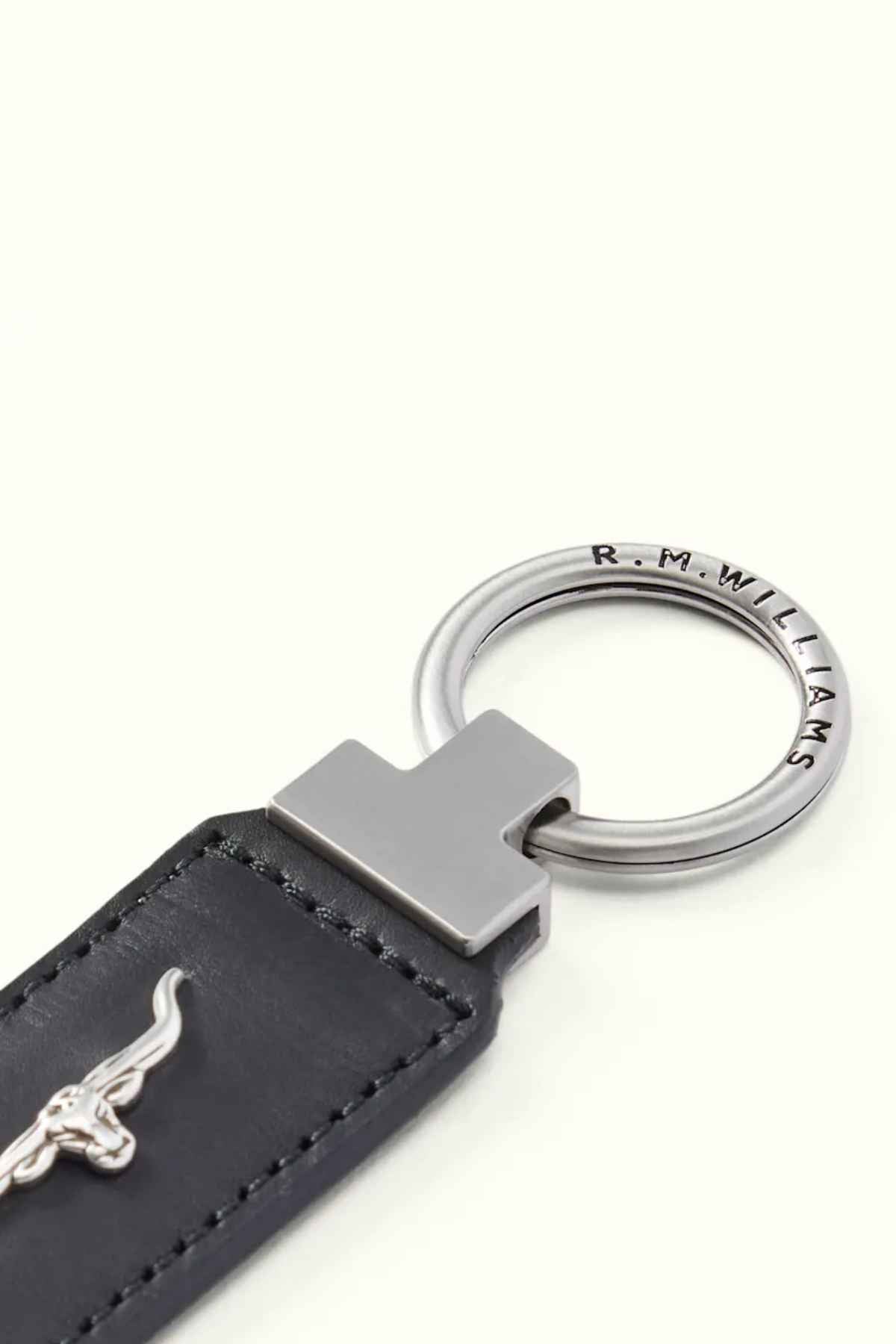 Close-up of the RM Williams Angaston Key Fob, showcasing a black leather tag adorned with a silver bull emblem that evokes the longhorn logo. The metal ring is engraved with "R.M. Williams" and is set against a plain, light background.