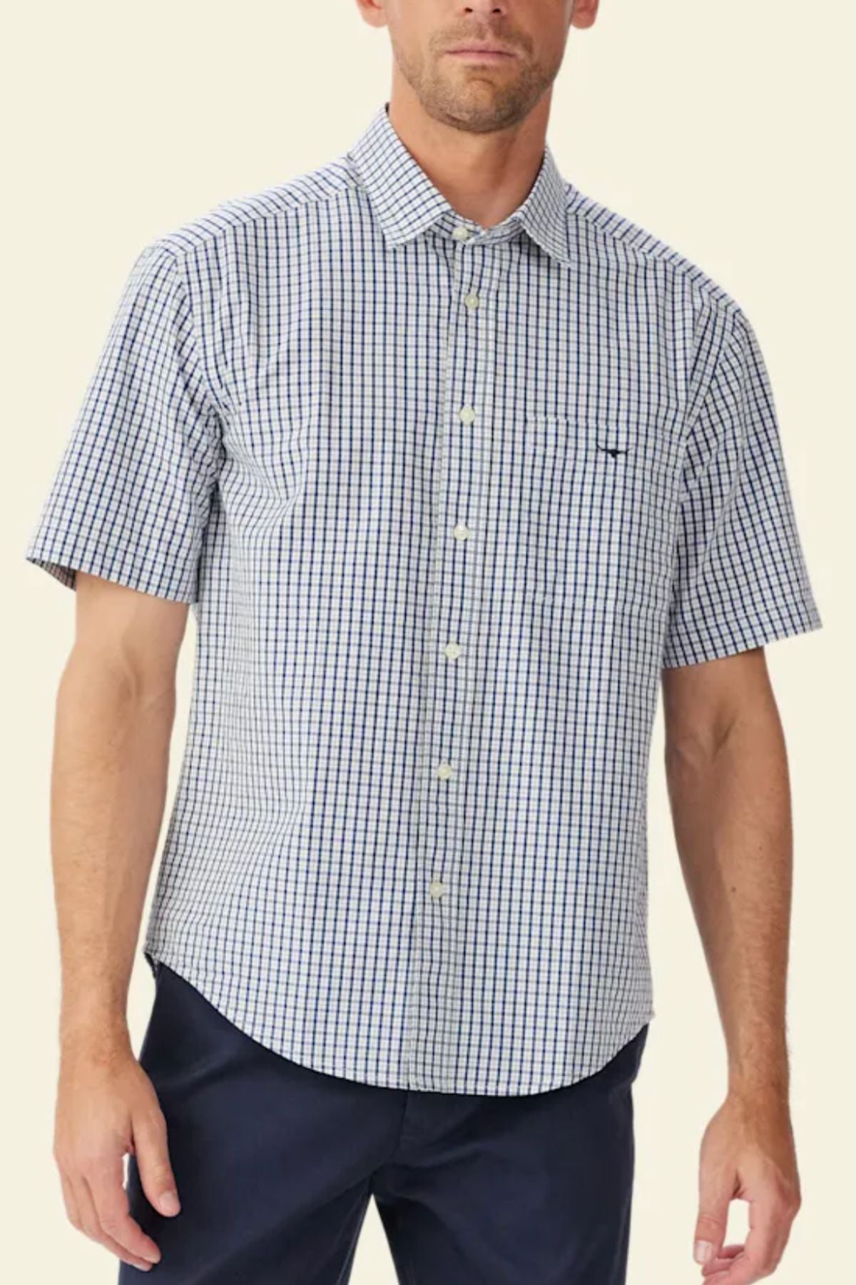 A man wears the RM Williams Augusta Poplin Short Sleeve Shirt, featuring a relaxed fit and blue and white checkered pattern, paired with dark pants, standing against a plain light background.