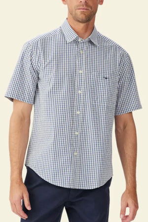 A man wears the RM Williams Augusta Poplin Short Sleeve Shirt, featuring a relaxed fit and blue and white checkered pattern, paired with dark pants, standing against a plain light background.