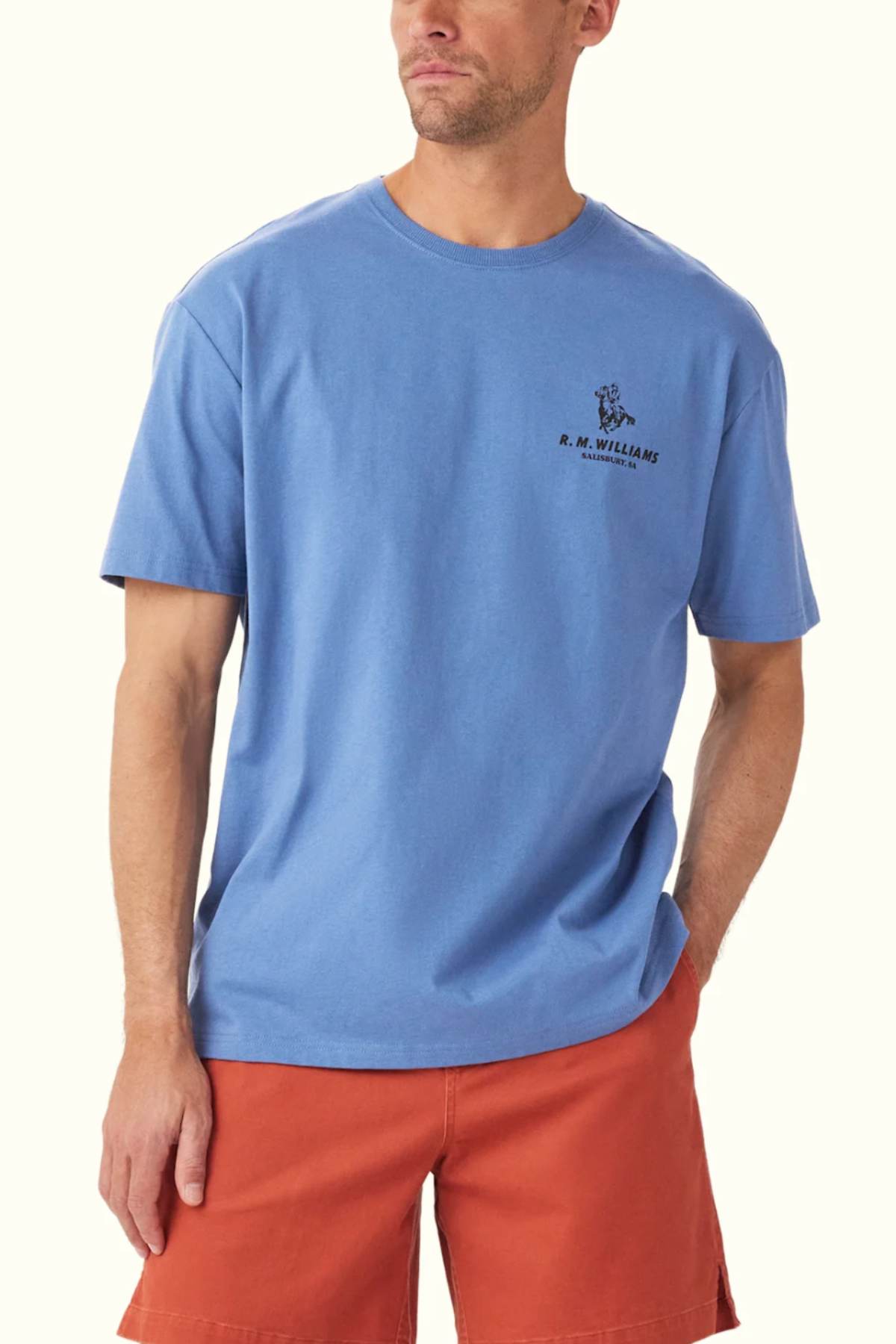 A person wearing a blue RM Williams Banner T-Shirt made from organic cotton, featuring a heritage logo design, paired with orange shorts. They are looking to the side with one hand in their pocket.
