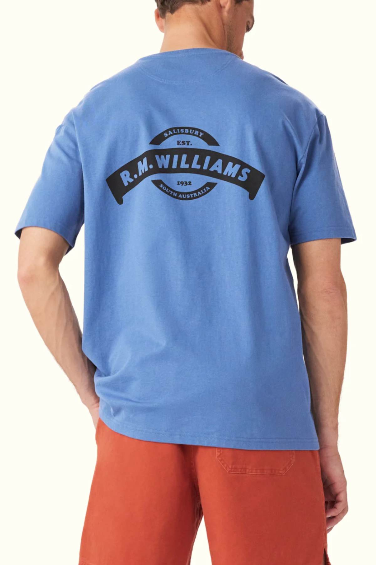 A man wearing a blue RM Williams Banner T-Shirt crafted from organic cotton displays an archive-inspired heritage logo design with the text "Salisbury Est. 1932 R.M. Williams, South Australia" on the back, paired with coral shorts. As he faces away from the camera, the shirt's design is prominently showcased.