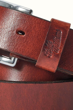 Close-up of the RM Williams Berwick Belt, featuring brown textured leather with a roller-style buckle and an embossed longhorn emblem on the leather keeper. The belt has a slightly worn finish for added character.