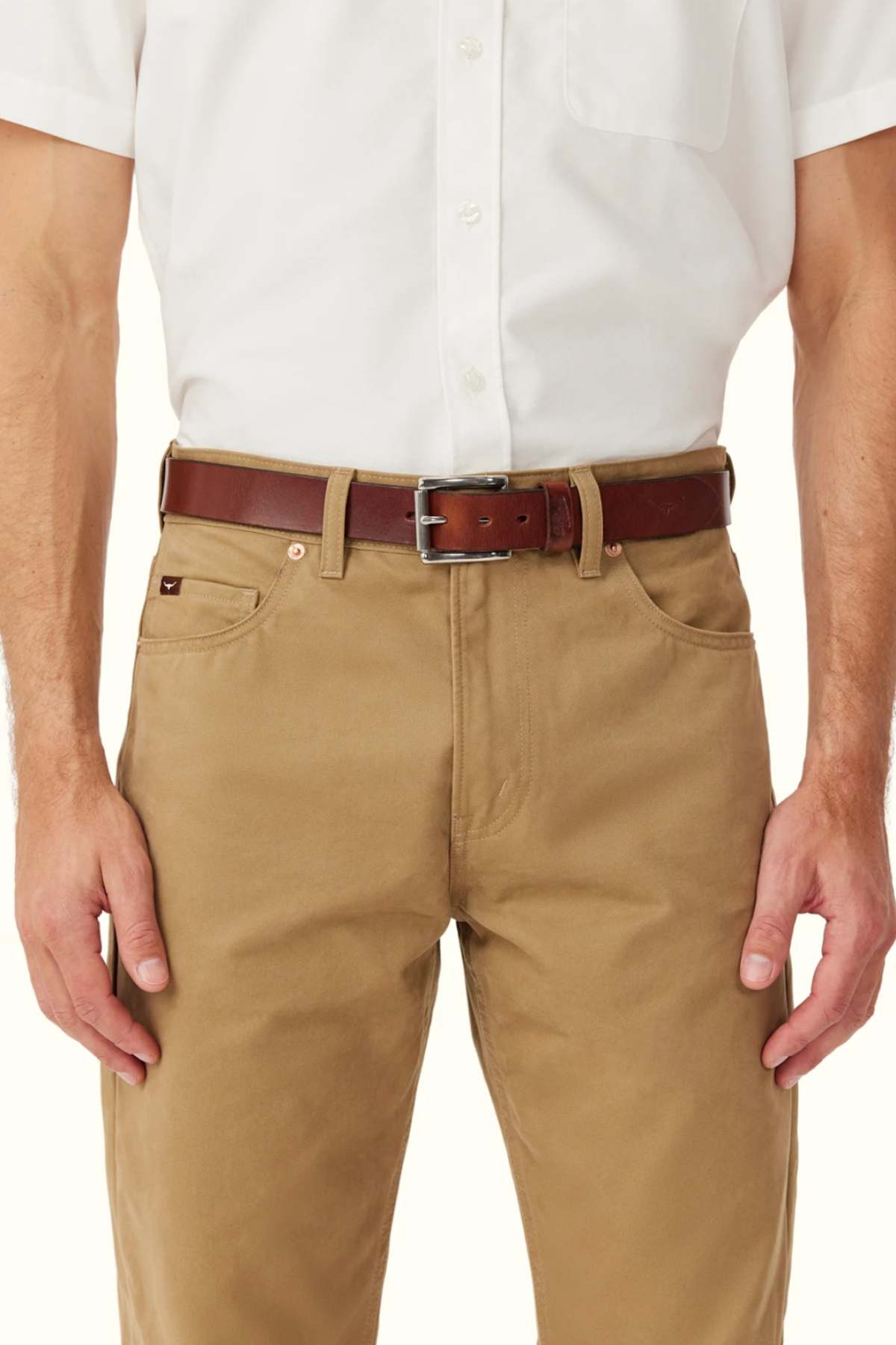 A person is shown from shoulders to mid-thigh wearing a white short-sleeve button-up shirt, tan pants, and the RM Williams Berwick Belt—a brown leather belt with a roller-style buckle.
