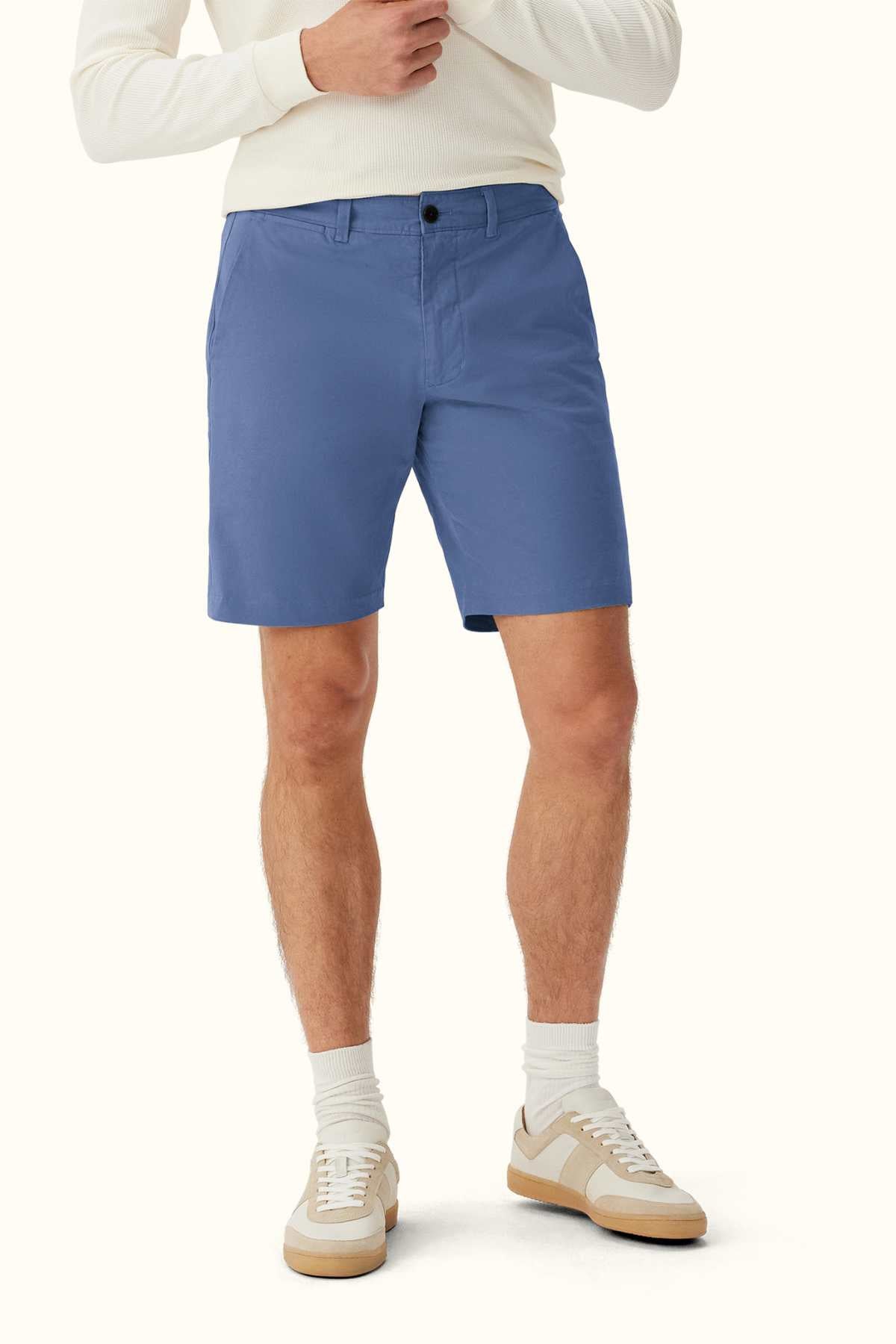 A person wears light blue RM Williams Berwick Chino Shorts paired with a white sweater, complemented by white socks and beige sneakers, standing against a plain background. Crafted from organic cotton for comfortable stretch, the ensemble presents a casual look ideal for warm weather.