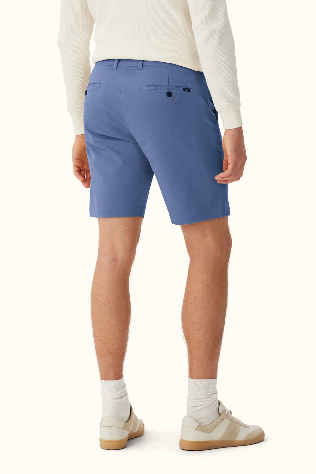 A person is seen wearing a white long-sleeve shirt and the RM Williams Berwick Chino Short, which are modern tailored shorts made from organic cotton. They are paired with white socks and beige sneakers, standing with their back facing the viewer. A plain, light-colored background emphasizes the simplicity and comfort stretch of their outfit.