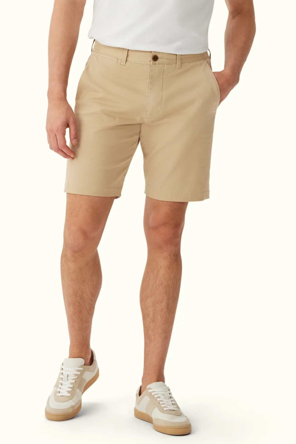 A person wears the RM Williams Berwick Chino Short, elegantly tailored and made from organic cotton, paired with a white shirt and white sneakers adorned with tan details. The shorts feature a button and pockets, offering both style and comfortable stretch as the individual stands against a plain white background.