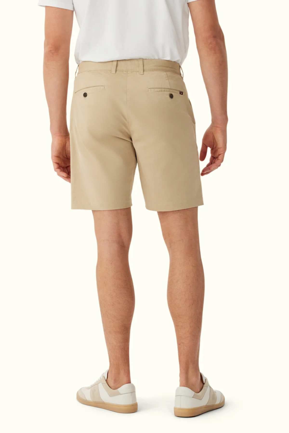 Back view of an individual wearing the RM Williams Berwick Chino Short in beige, crafted from organic cotton, paired with a crisp white shirt. They stand with legs slightly apart, highlighting the buttoned back pockets. White sneakers with beige soles complete the look, adding casual elegance.
