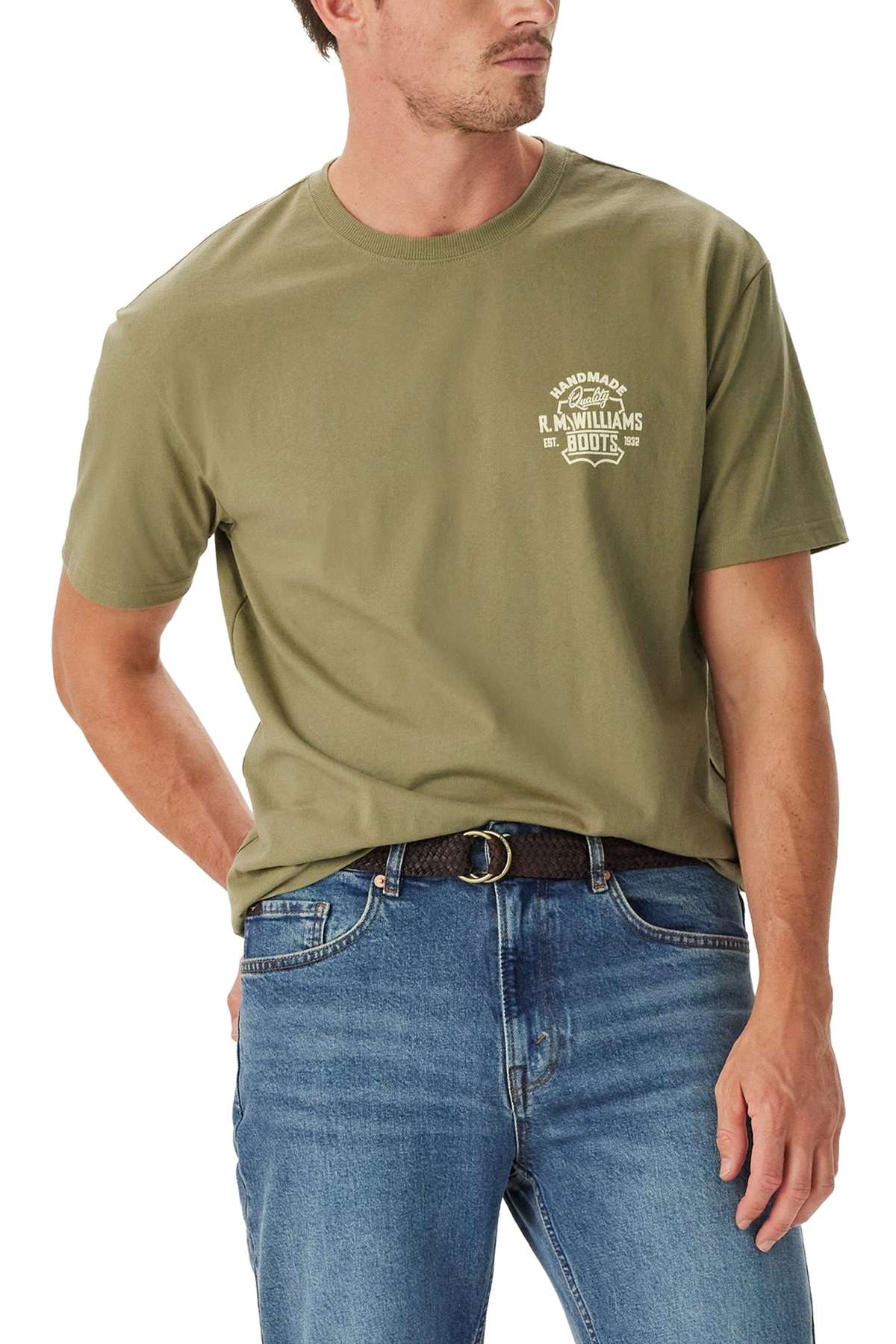A man wearing the RM Williams Boot T-Shirt in plain olive green, featuring a small white logo on the left chest, paired with blue jeans and standing with one hand in his pocket, looking to the side.