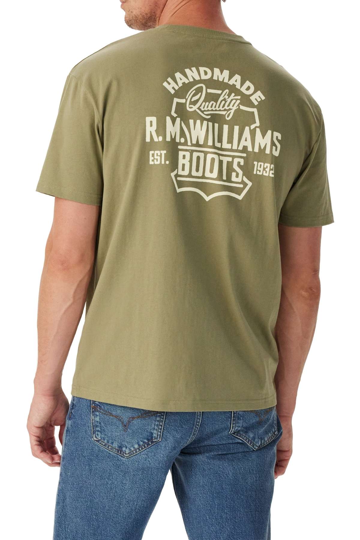 A man stands with his back to the camera, wearing the RM Williams Boot T-Shirt in olive green, featuring white "Handmade Quality R.M. Williams Boots Est. 1932" text on the back, paired with blue jeans.