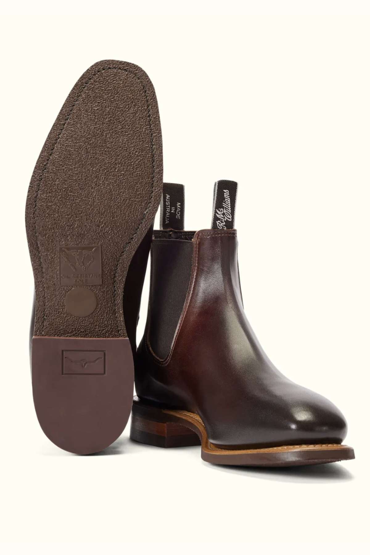 A pair of RM Williams Burnished Comfort Craftsman Boots in dark brown leather; one boot stands upright highlighting its side and elastic panel, while the other lies on its side showing the textured rubber sole. Made in Australia.