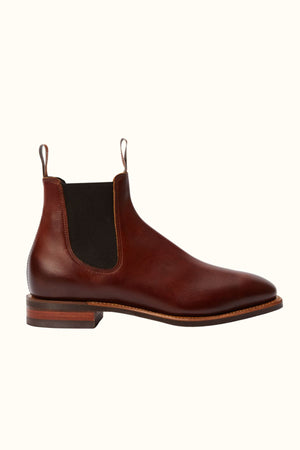 The RM Williams Burnished Comfort Craftsman Boot is a brown hand-burnished leather Chelsea boot with a rounded toe, black elastic side panel, front and back pull tabs, and a low stacked heel, shown on a plain light background.