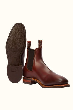 The RM Williams Burnished Comfort Craftsman Boot features brown hand-burnished leather, chisel toes, elastic side panels, and pull tabs; one boot stands upright while the other displays its textured sole and heel.