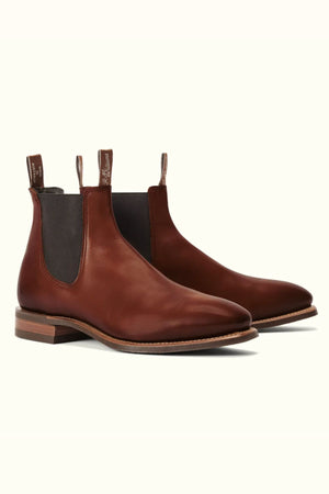 The RM Williams Burnished Comfort Craftsman Boot features brown hand-burnished leather, elastic side panels, pull tabs, a chisel toe, and low wooden heels, shown against a plain light background.