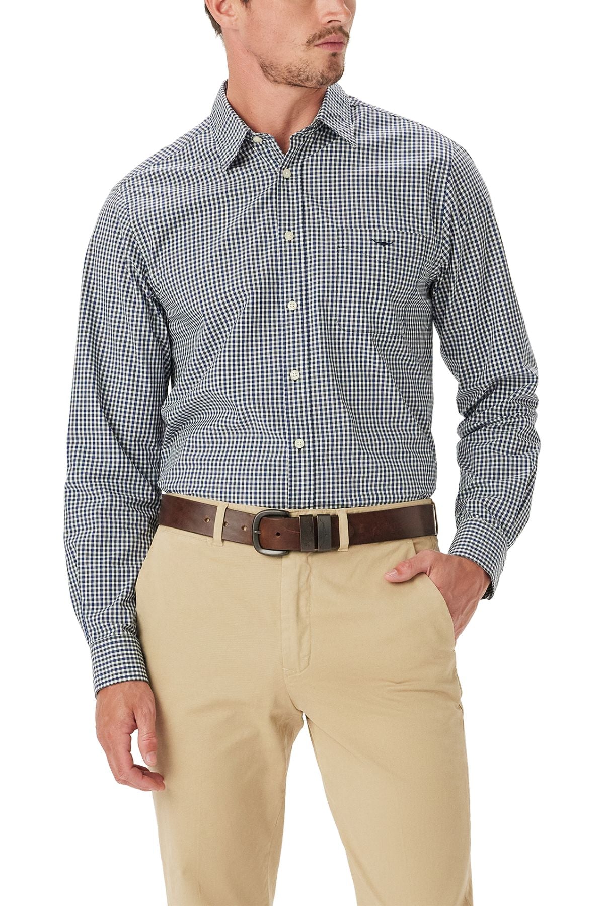 A man wearing the RM Williams Classic Poplin Check Shirt in blue and white, tucked into beige pants with a brown belt, stands with one hand in his pocket.