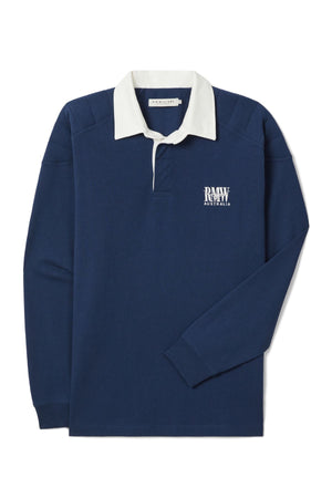 The RM Williams Classic R.M.W Rugby is a navy blue, long-sleeve Australian cotton shirt with a white collar, partial button placket, and “RAW AUSTRALIA” text plus logo embroidered in white on the left chest.