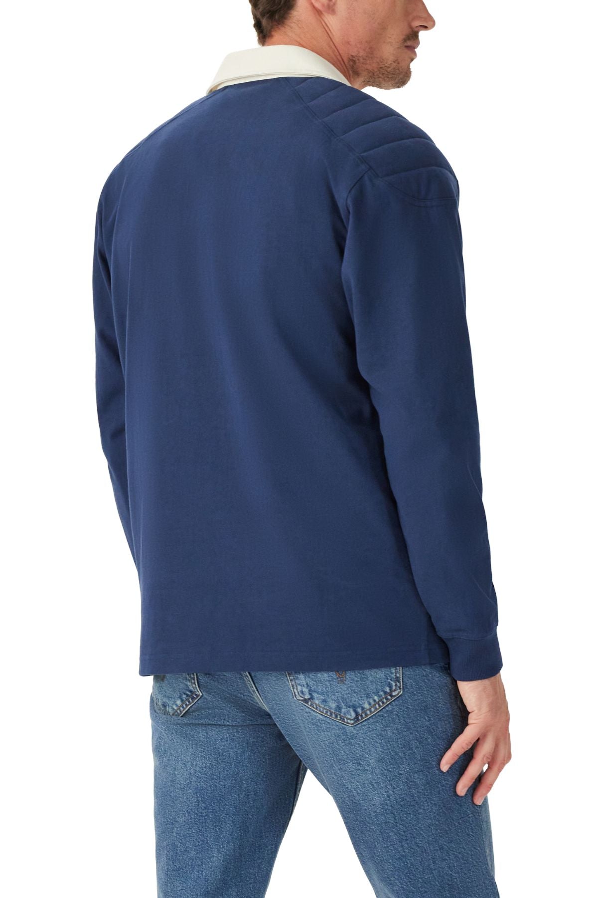 A man faces away, wearing the RM Williams Classic R.M.W Rugby—a blue heavyweight Australian cotton jersey with a white collar—paired with light blue jeans. Only his upper body and profile are partly visible.