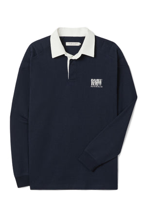 The RM Williams Classic R.M.W Rugby shirt features heavyweight Australian cotton in navy blue, a contrasting white collar, and a white "RAW AUSTRALIA" logo on the left chest. It’s shown laid flat with the right sleeve crossed over.