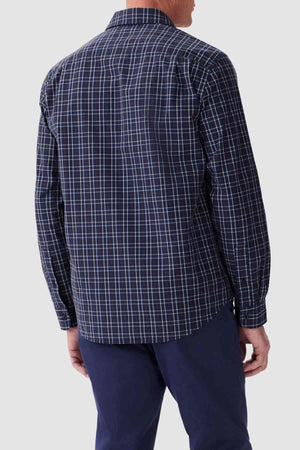 A person wearing an RM Williams Classic Shirt, featuring a navy blue and white checkered pattern, is shown from behind. The long-sleeve shirt, made of organic cotton with a regular fit and spread collar, has its sleeves down. They are also dressed in dark pants against a plain, light-colored background.