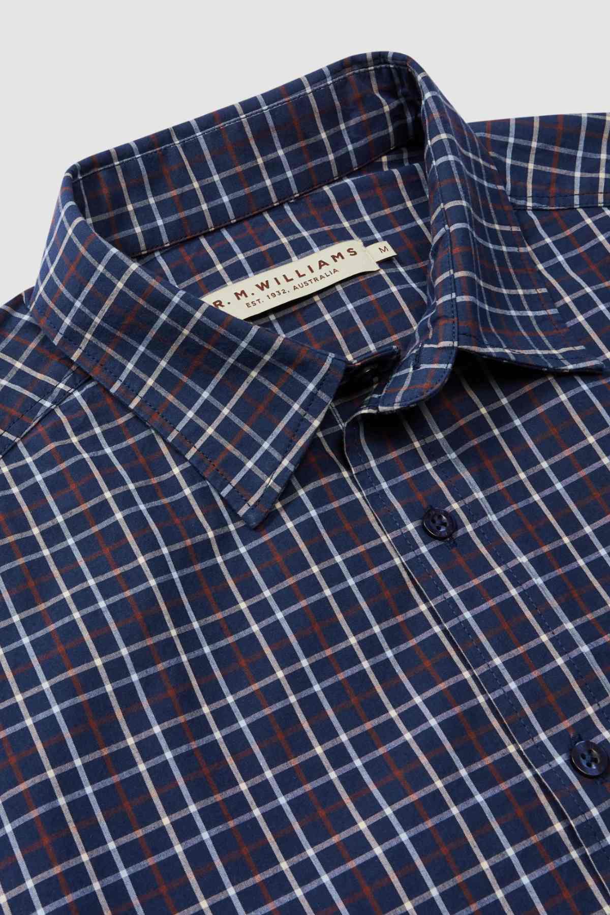 Here's a close-up of the RM Williams Classic Shirt, a blue, red, and white plaid button-up with a collar. This smart casual shirt features a label from "R.M. Williams" at the collar and is available in size medium (M). Made from organic cotton, the fabric boasts a crisp appearance with buttons running down the front.