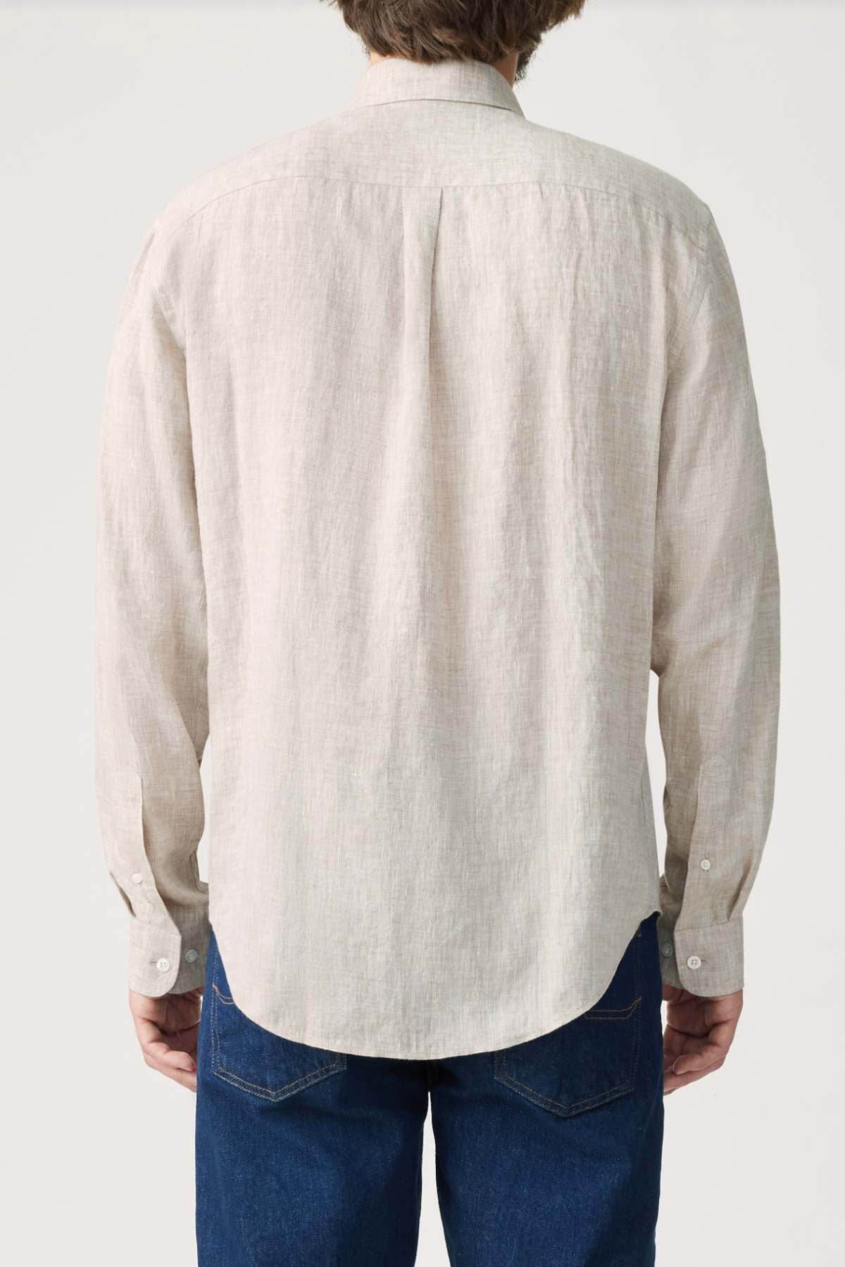 A person is seen from behind, wearing a light beige RM Williams Classic Shirt with a button down collar and blue jeans, standing against a plain light background.