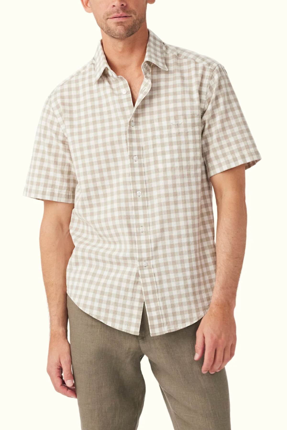 A man dressed in a RM Williams Classic Short Sleeve Shirt, featuring a beige and white gingham pattern, stands against a plain backdrop. This French linen shirt is fully buttoned up, and he pairs it with olive green pants. His head is slightly tilted, displaying a neutral expression.