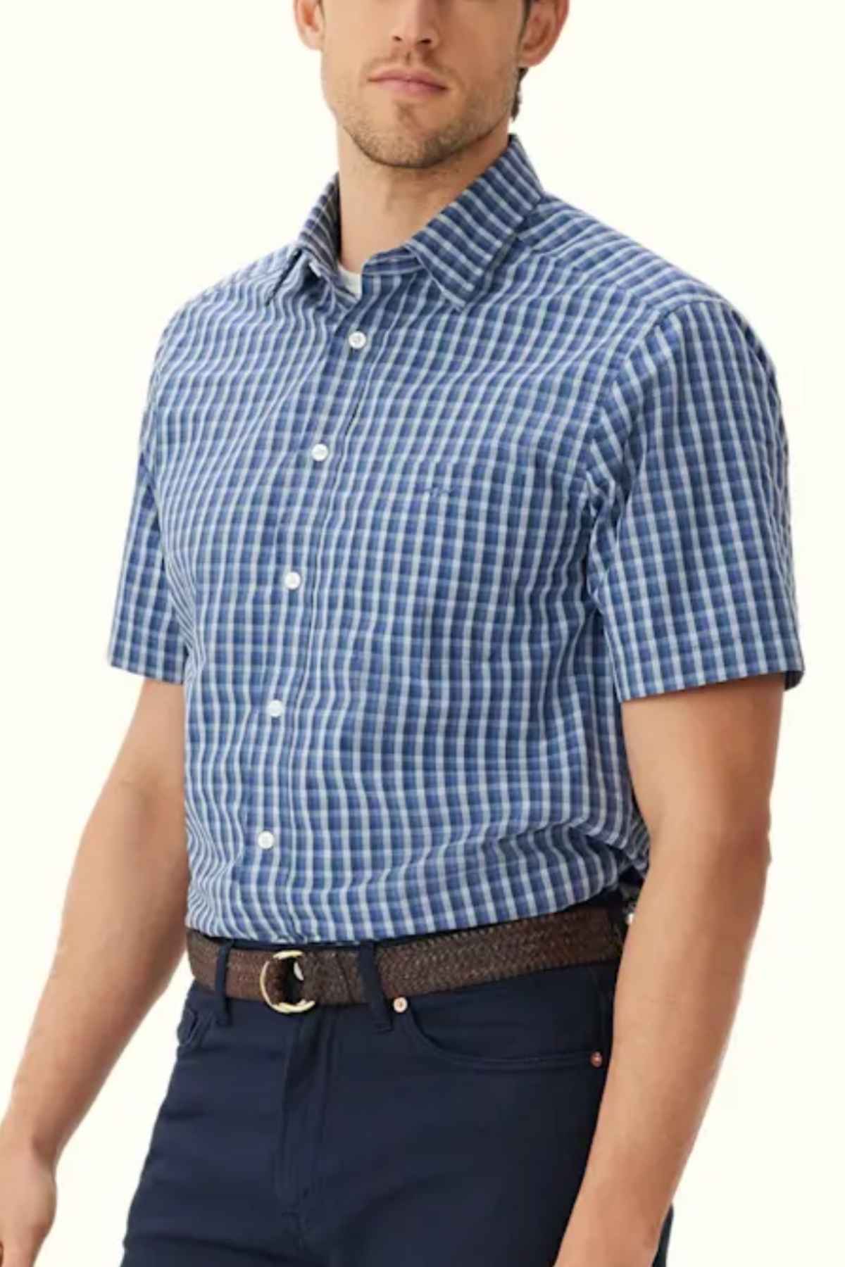 A man wearing an RM Williams Classic Short Sleeve Shirt, featuring a blue and white checkered pattern, regular fit, and classic collar with white buttons. The shirt is tucked into dark blue pants secured with a brown belt. The background is plain and light-colored.