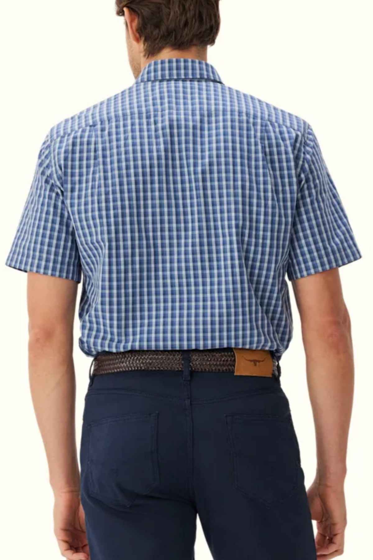 A man is standing with his back to the camera, dressed in a regular fit RM Williams Classic Short Sleeve Shirt featuring a blue and white checkered pattern made of cotton poplin. He wears dark blue pants and a braided brown belt, with his hands resting casually at his sides.