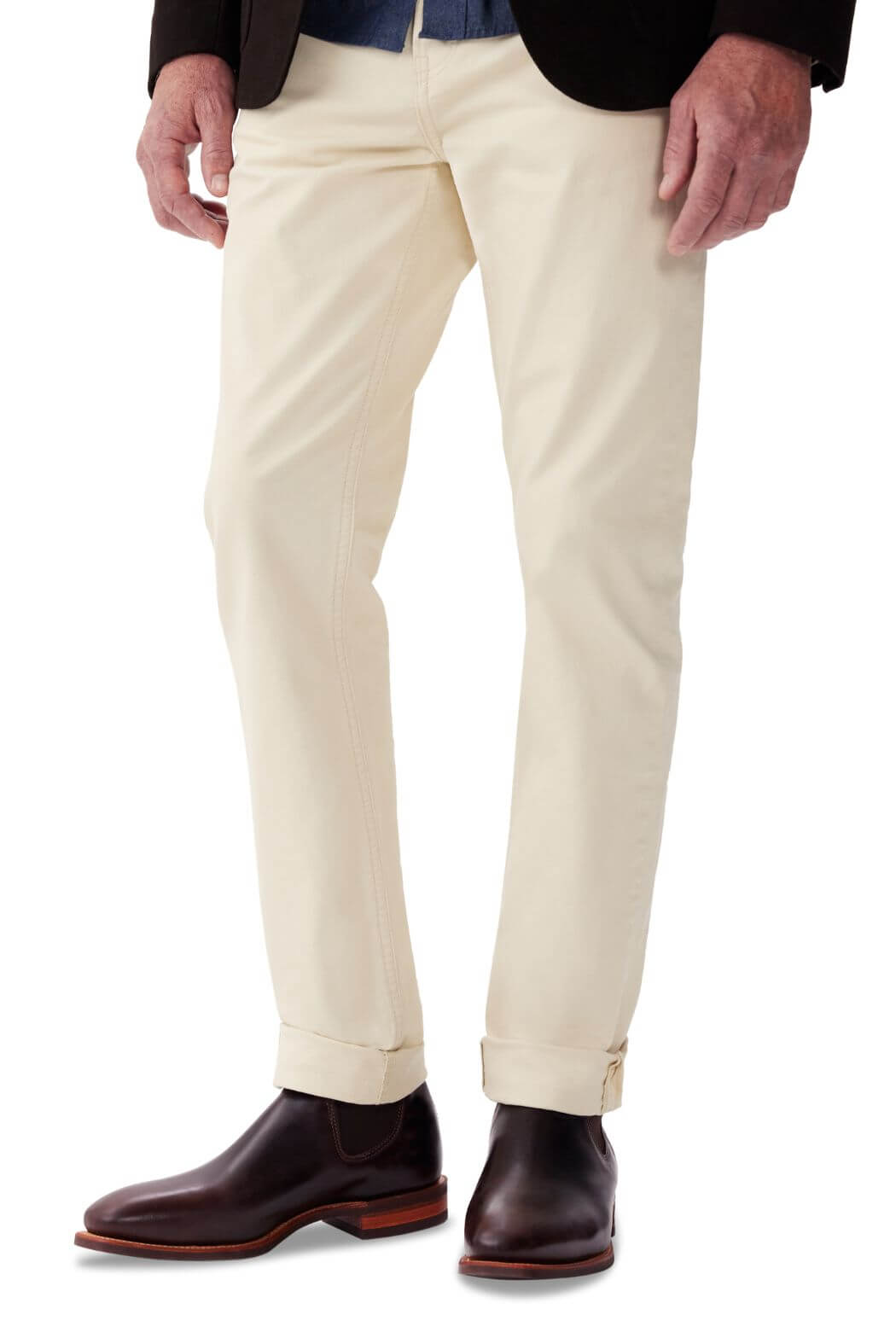 A man wearing beige pants and dark brown RM Williams Comfort Craftsman Boots made in Australia stands with one hand by his side and the other partially visible. The pants are slightly rolled up at the hem, and he is dressed in a dark blazer with a blue shirt underneath.