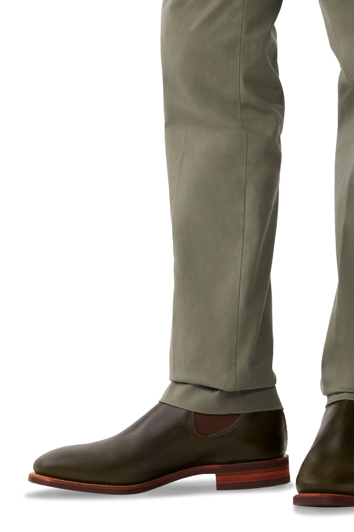A person stands against a white background, wearing olive green pants and RM Williams Comfort Craftsman Boots in pull-up leather with reddish-brown soles; only their lower legs and feet are visible.