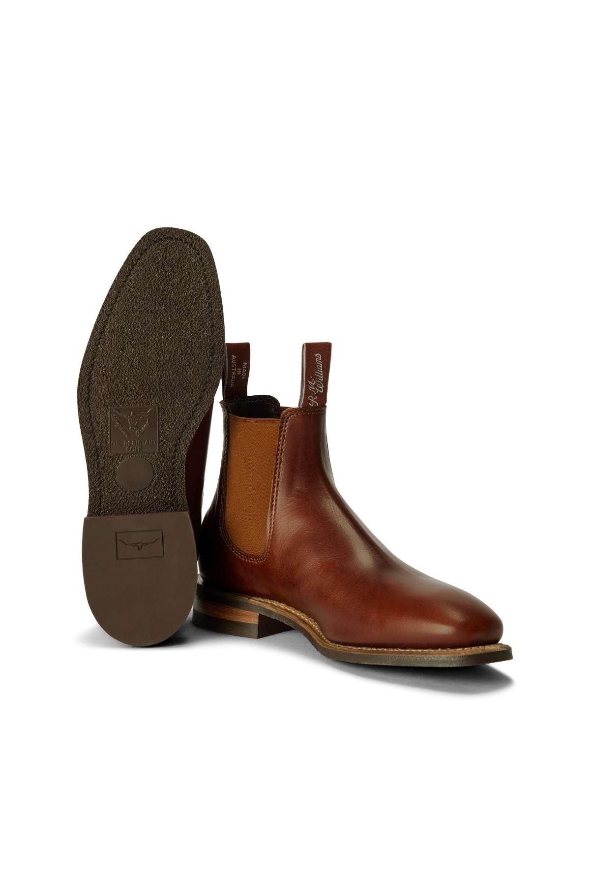 A pair of RM Williams Comfort Craftsman Boots in brown leather, featuring elastic side panels, pull tabs, and a low heel; one stands upright while the other displays its textured sole.