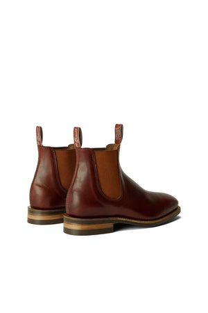 An RM Williams Comfort Craftsman Boot in brown leather, featuring elastic side panels, pull tabs, and stacked wooden heels, is displayed on a white background facing slightly away.