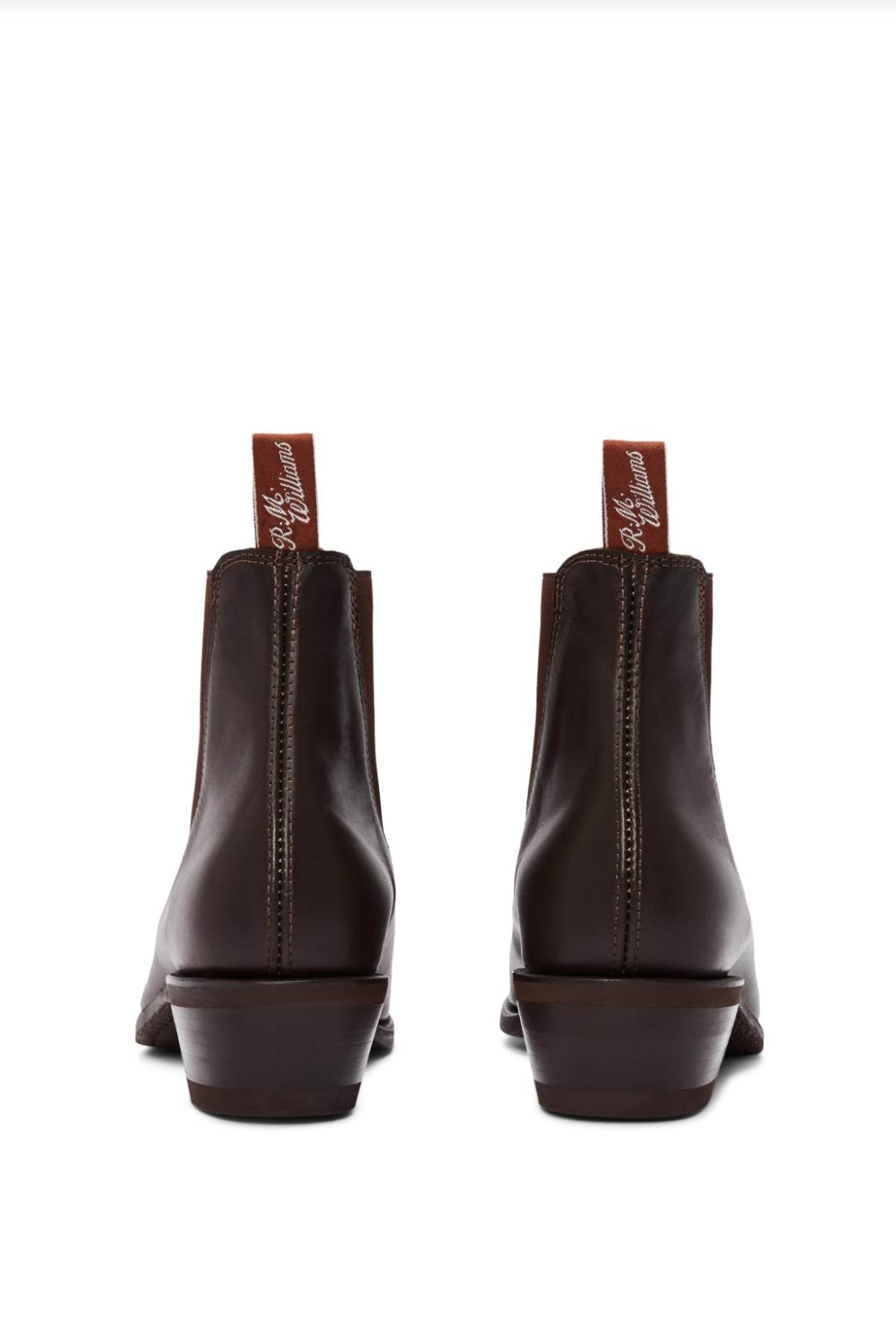 The RM Williams Comfort Lady Yearling Rubber Sole Boots are pictured from the back, featuring dark brown leather in the classic Yearling boot style. The boots have reddish-brown pull tabs with white cursive text and exhibit a slight heel along with visible stitching, elegantly displayed against a plain white background.