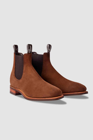 The RM Williams Comfort Suede Craftsman Boot is a brown suede Chelsea boot with dark elastic sides, front and back pull tabs, and low wooden heels, made in Australia and shown on a light gray background.