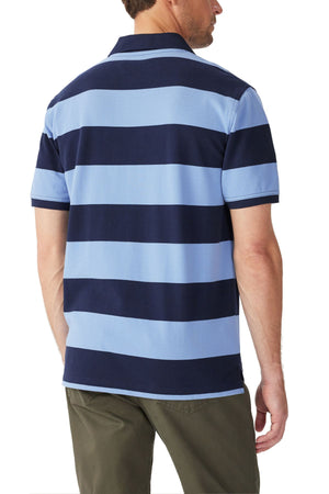 A man is shown from the back wearing the RM Williams Craddle Polo, a short-sleeve collared rugby shirt with wide navy and light blue stripes in organic cotton, paired with olive green pants.