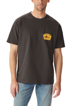 A man wears the RM Williams Craftsman T-Shirt—a loose-fit black short sleeve tee with a small yellow logo on the left chest—paired with light blue jeans, standing against a white background.