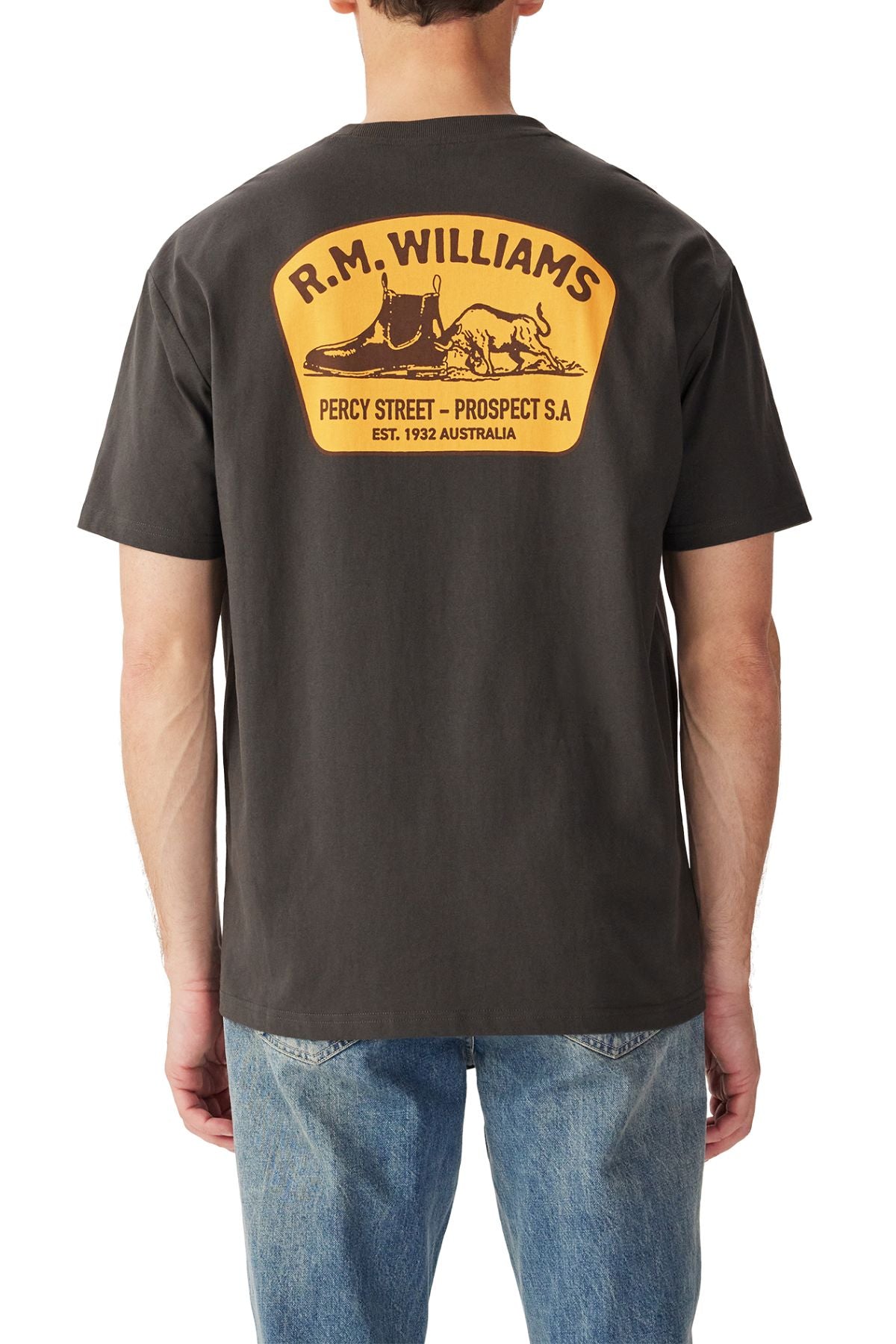 A man wears the black RM Williams Craftsman T-Shirt with a large yellow and brown boots and hat "R.M. Williams" print on the back, featuring "Percy Street - Prospect S.A. EST. 1932 Australia," paired with blue jeans.